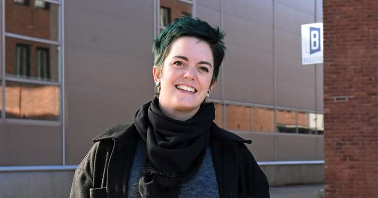 En kvinne med mørkt, grønt hår og mørke klær står utendørs foran en bygning med vinduer og rødt murverk.