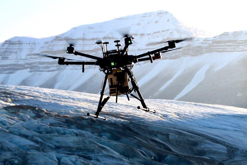 Drone i luften med snøkledde fjell i bakgrunnen.
