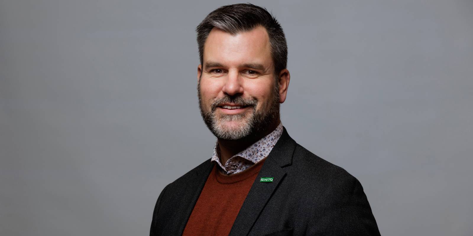 En mann med kort, mørkt hår har på seg dressjakke, rød genser og skjorte med mønster mot grå bakgrunn.