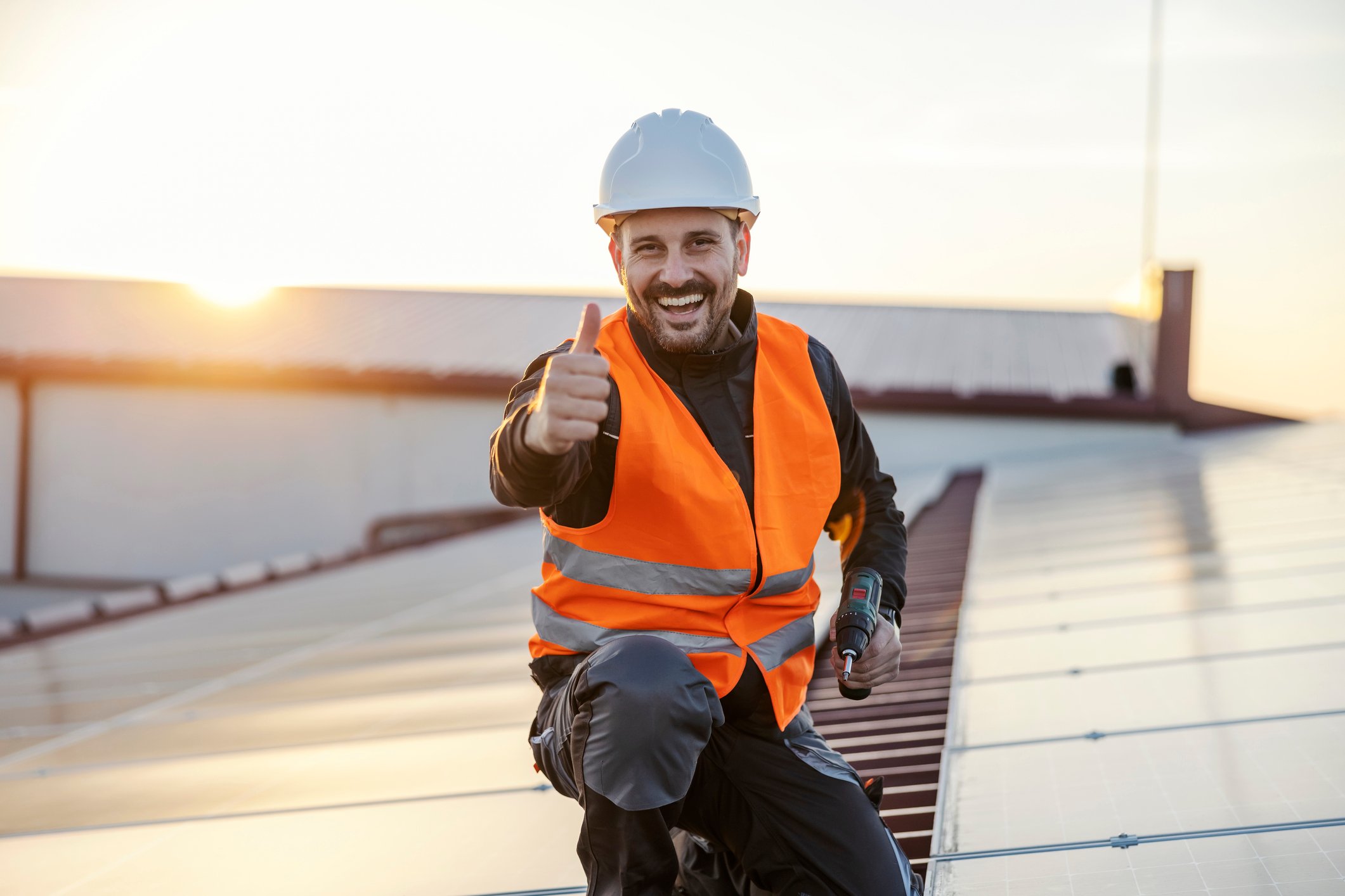 Smilende håndverker iført hvit hjelm og oransje refleksvest gir tommel opp mens han står på et tak med solcellepaneler.