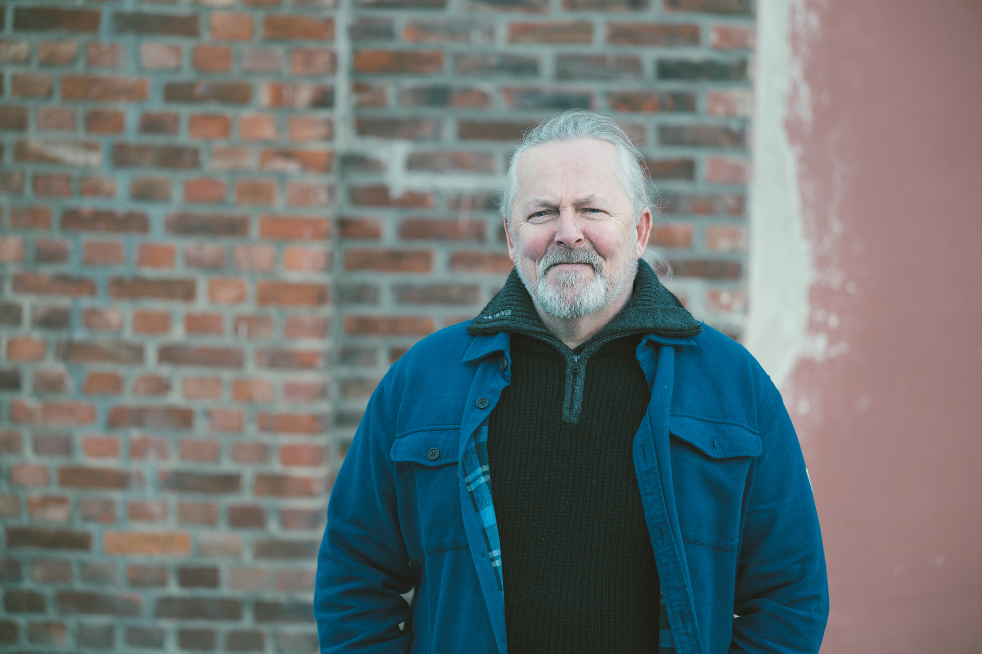 Mann med grått hår og skjerf står foran et murbygg. Har på seg svart genser og blå jakke.