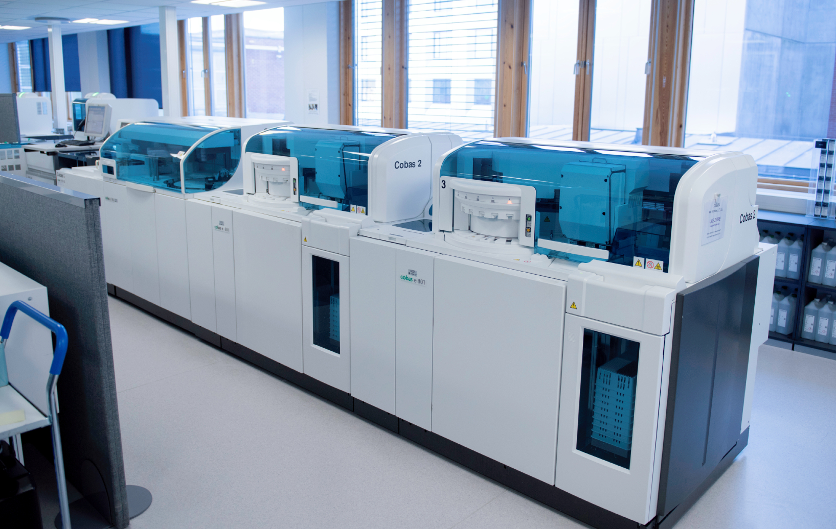 Bildet viser et moderne, lyst laboratorium med store maskiner for blodprøveanalyse. Maskinene er hvite med blått lokk.