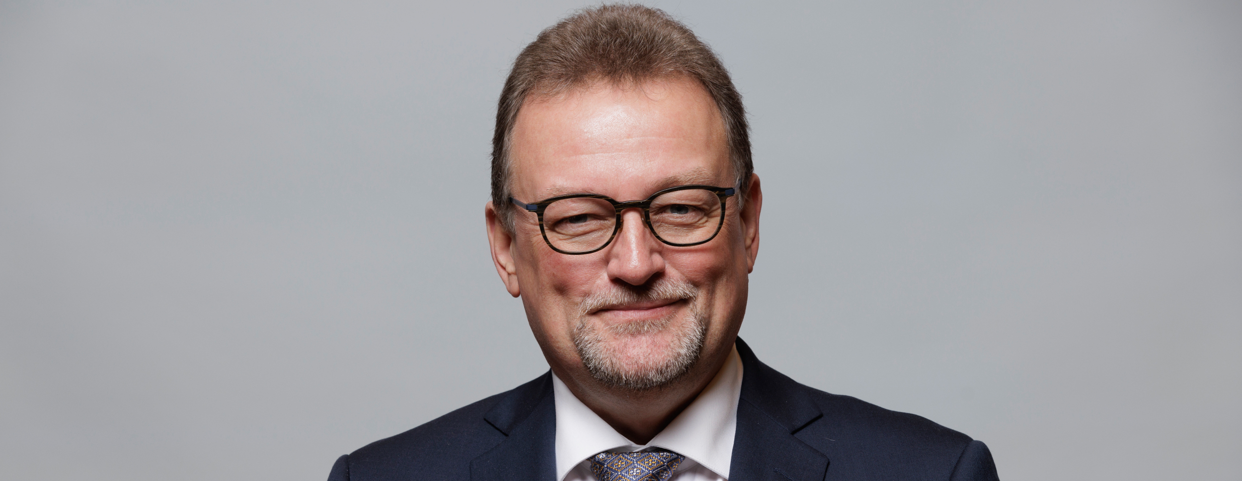 En smilende mann i dress og briller ser rett i kamera. Han har kort, brunt hår og skjeggstubb, og utstråler trygghet og vennlighet.