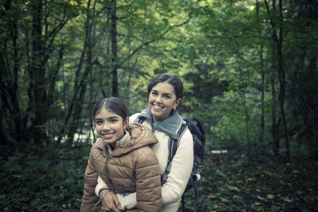 En kvinne og en jente med ryggsekk smiler og holder rundt hverandre mens de er ute på tur i en grønn, frodig skog.