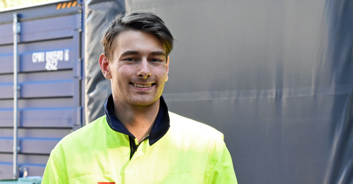 En person iført gul refleksjakke står foran en container på et industriområde.