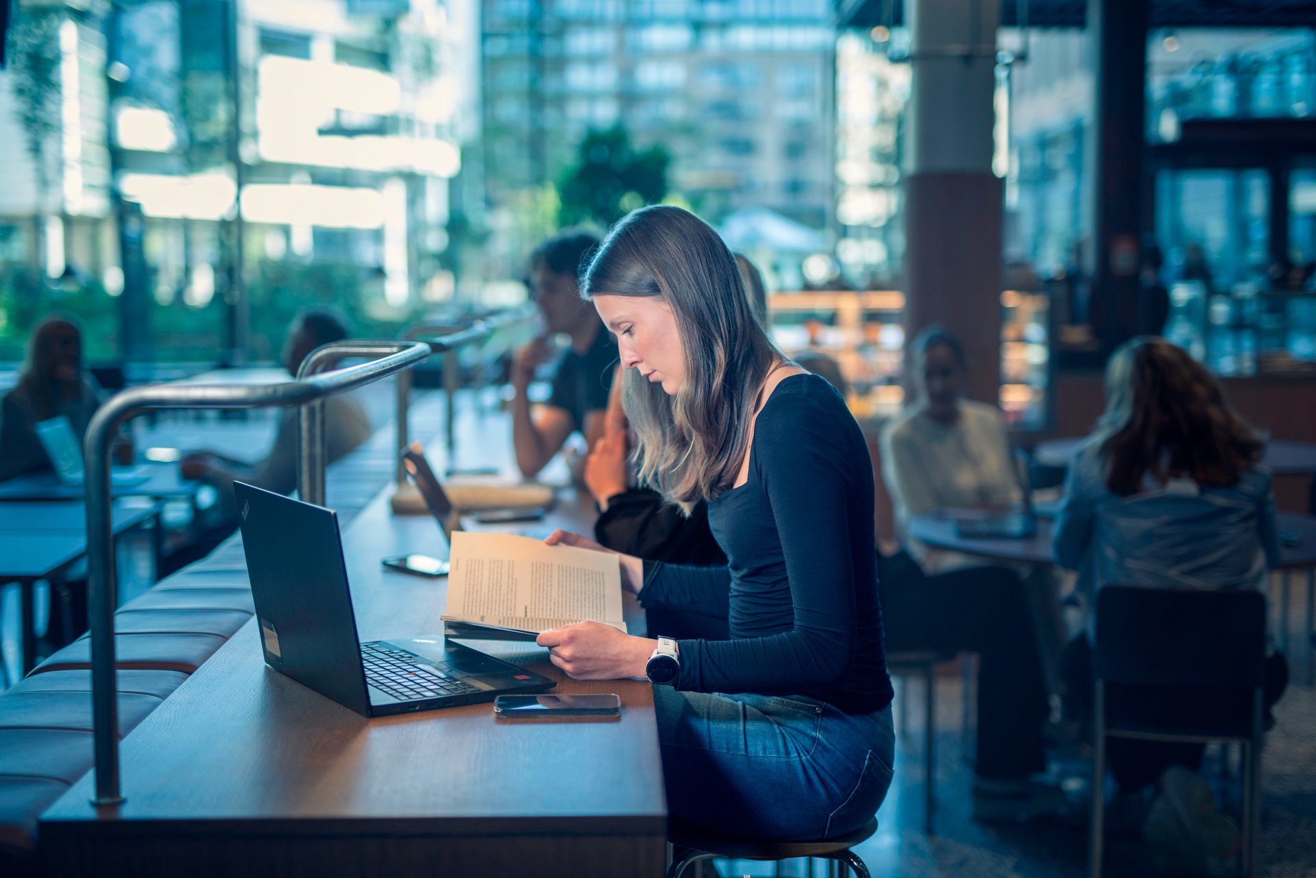 Ung kvinne studerer ved et bord i et moderne, lyst bibliotek, omgitt av bøker, laptop og andre studenter.