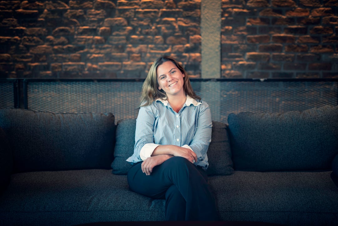 En kvinne med lyst hår sitter avslappet og smilende i en mørk sofa mot en vegg av stein eller murstein.