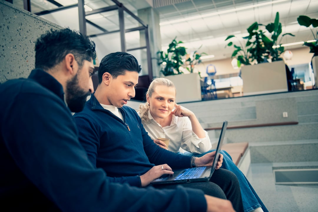 Tre personer sitter sammen på en trapp innendørs, ser på en bærbar PC og smiler.