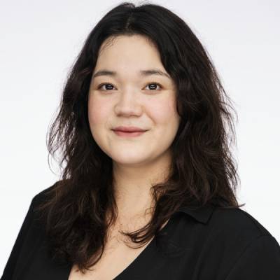 Person med mellomlangt, mørkt og litt bølgete hår, iført mørk skjorte, fotografert mot lys bakgrunn.