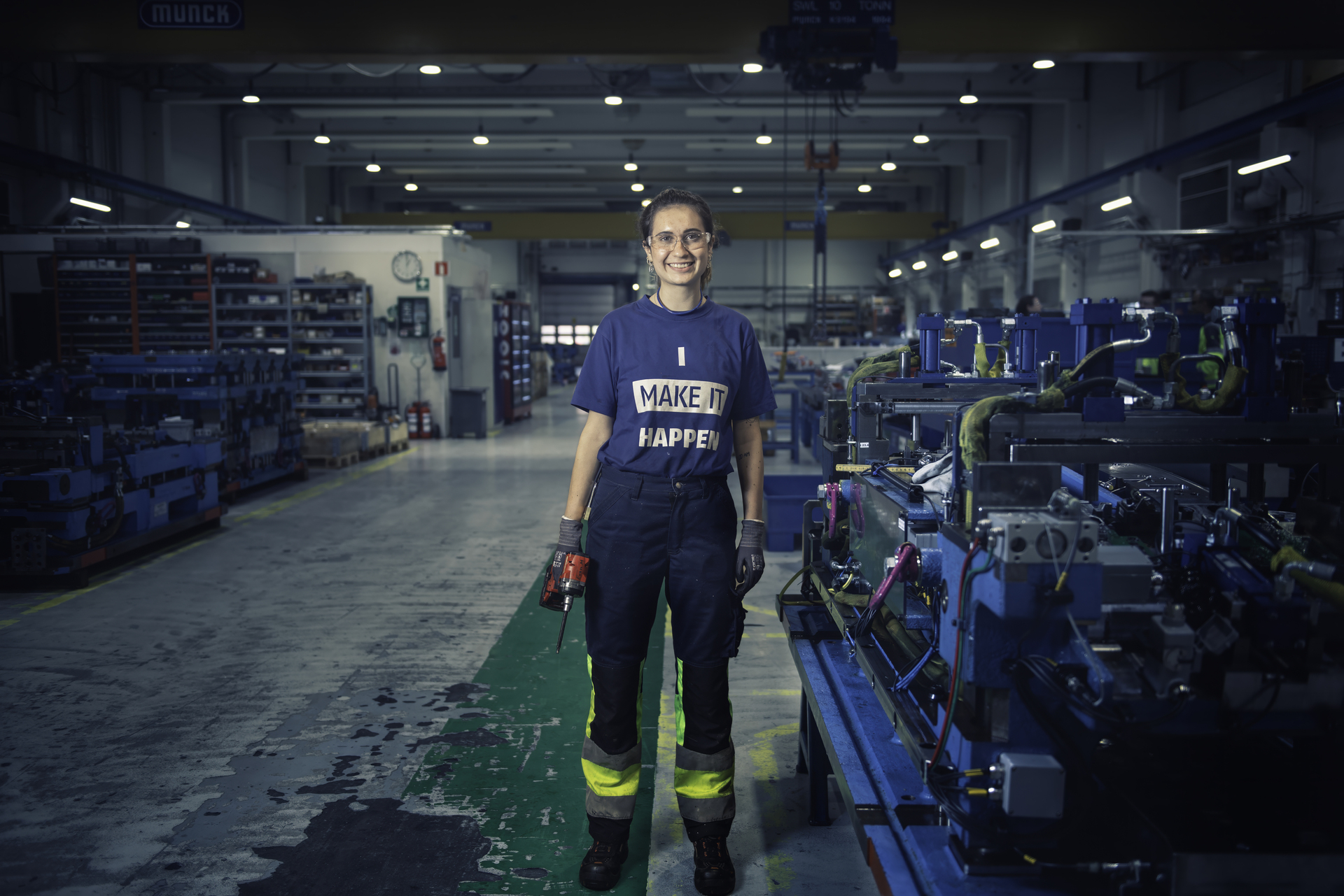 A young female engineer stands smiling at her workplace with industrial machinery in the background, her T-shirt says "I make it happen".