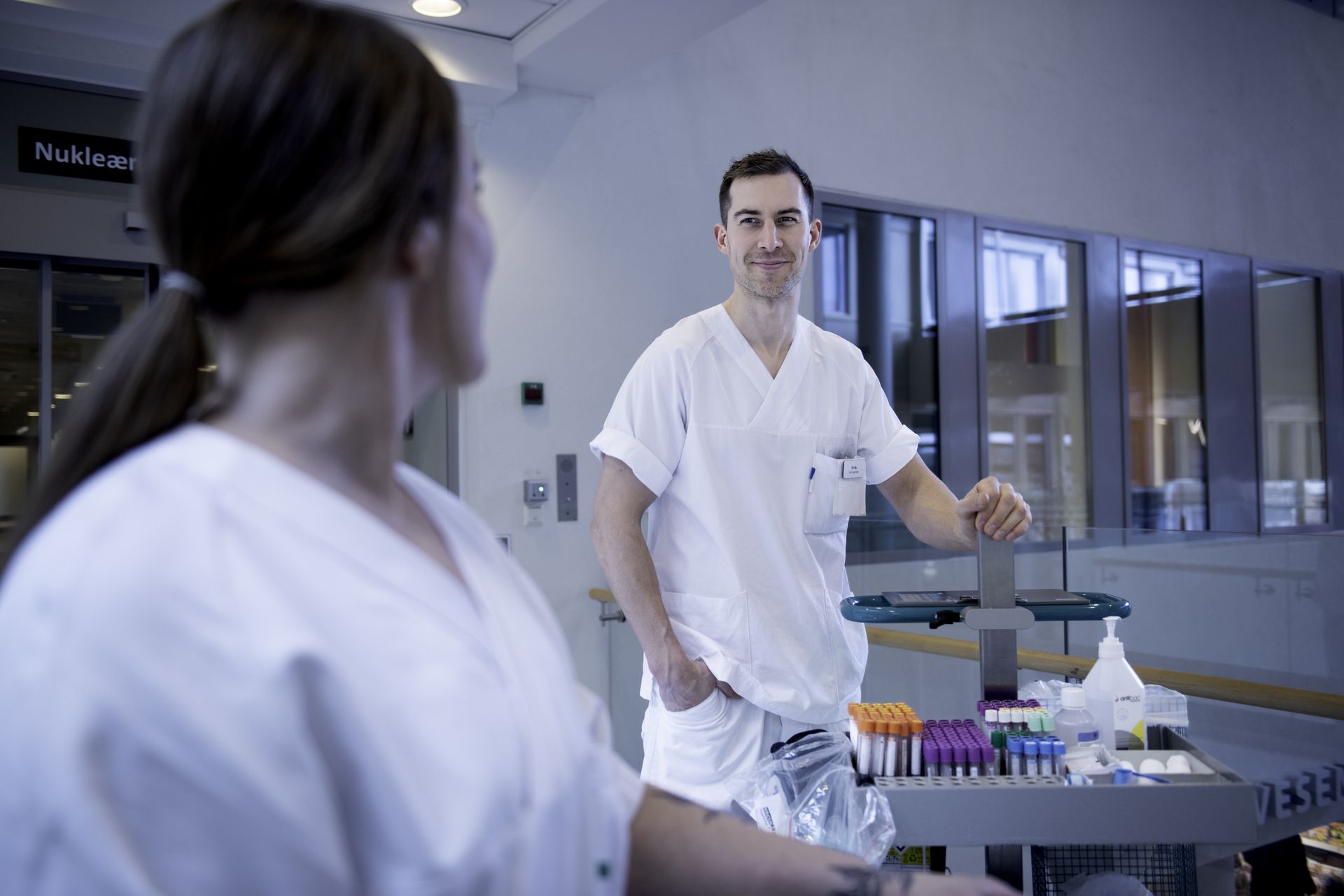 To uniformerte bioingeniører helsearbeider smiler til hverandre på sykehuset med en tralle med prøvetakingsutstyr 