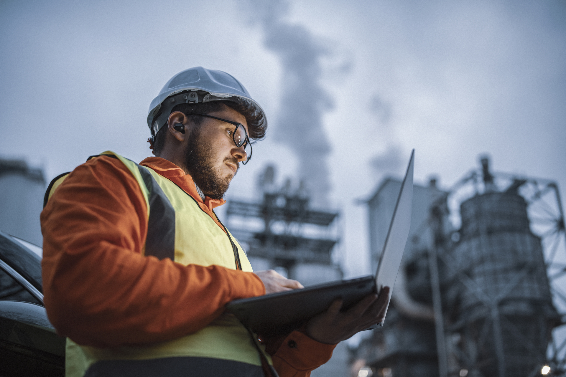 A serious engineer in protective equipment uses a laptop in an industrial environment, symbolizing the importance of good working conditions, safety and collective agreements in industry.