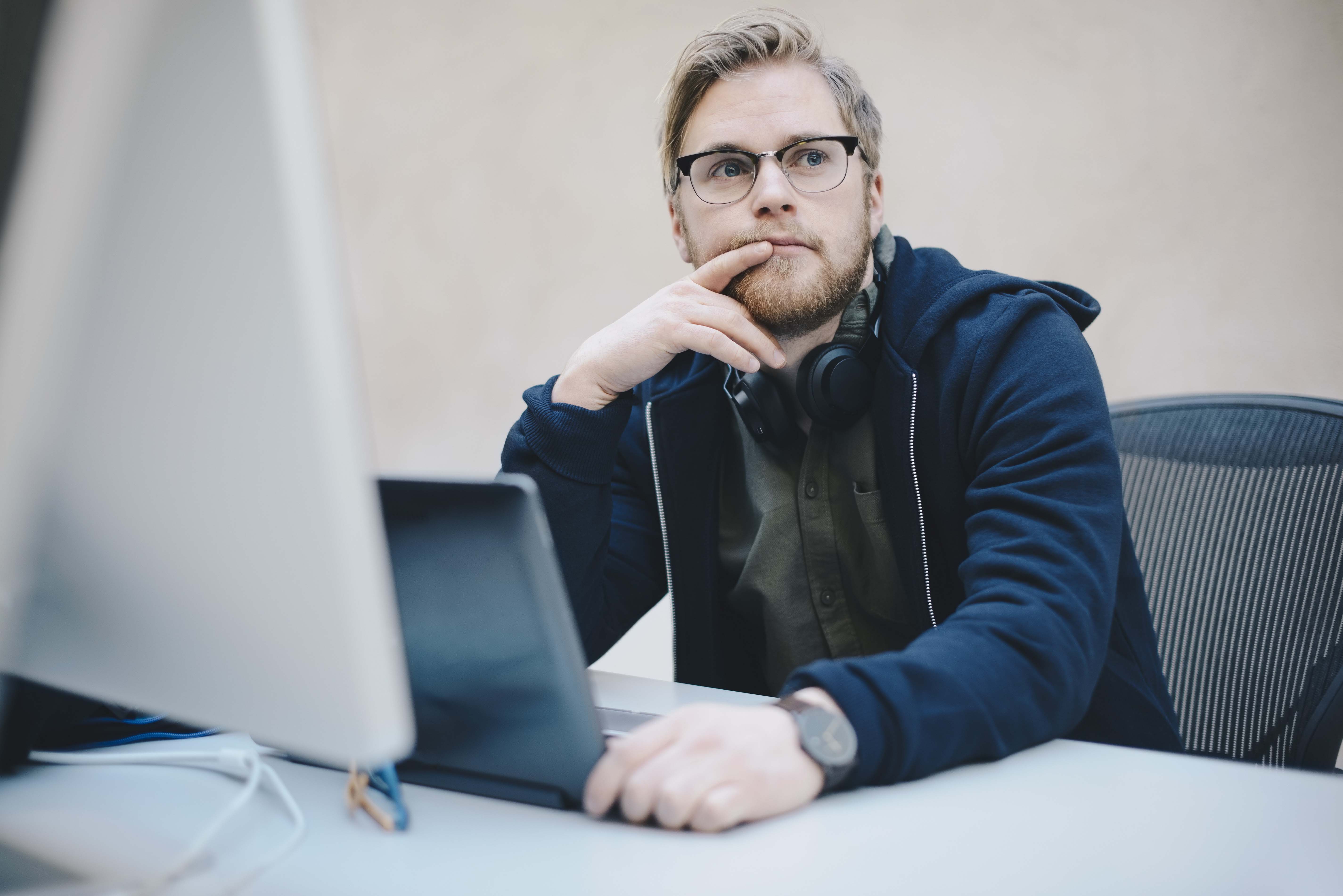A man with a beard and glasses sits at a computer with headphones around his neck, and looks over thoughtfully. The image may symbolize reflection or planning in connection with layoffs.