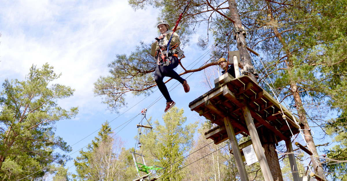 En dame som henger i en zipline, omgitt av grønne trær og en blå himmel.