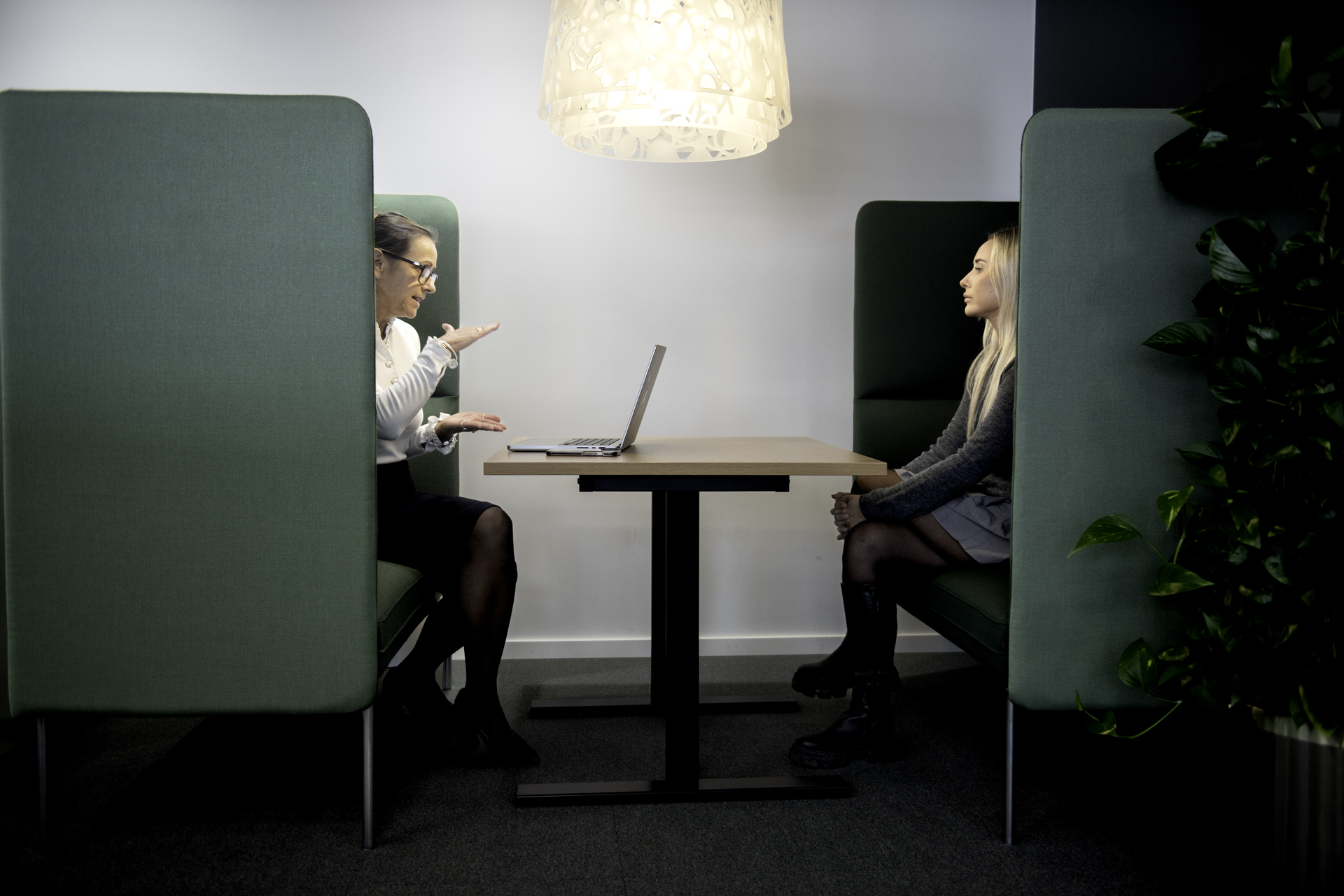 To personer sitter i et moderne møterom med grønne avskjermede båser, et bord mellom seg og en lampe over.