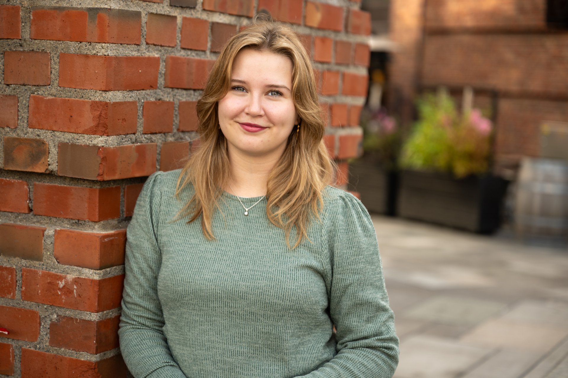 Ung dame ser i kameraet og lener seg mot en mursteinsvegg
