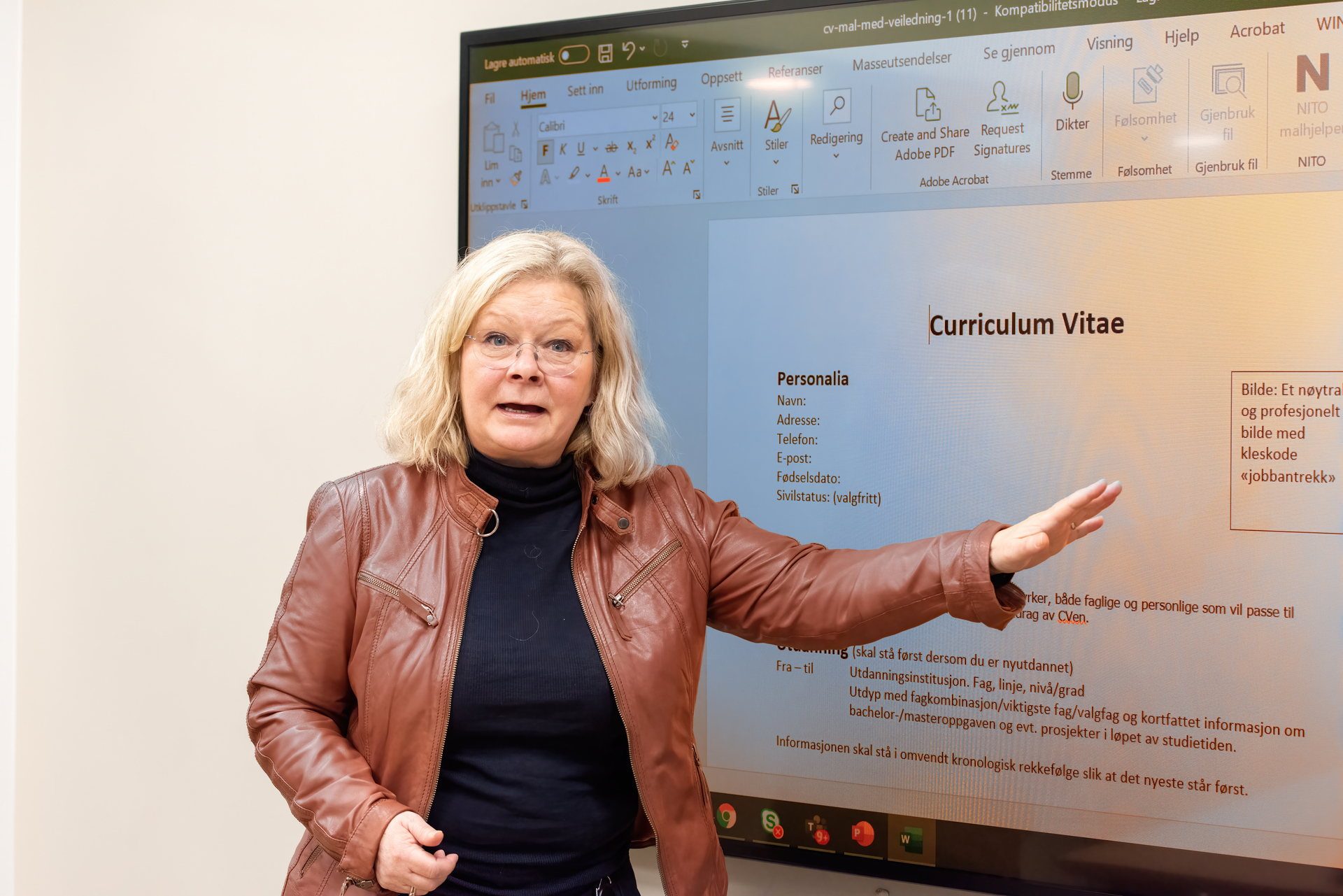 Image of woman with blond hair, glasses and brown leather jacket standing in front of a screen wall
